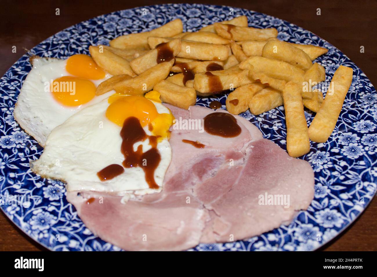 Prosciutto uova e patatine su un piatto di porcelano rotondo blu e bianco con motivi floreali su un tavolo di legno scuro. Uova di tuorlo doppio e di tuorlo singolo con salsa marrone Foto Stock