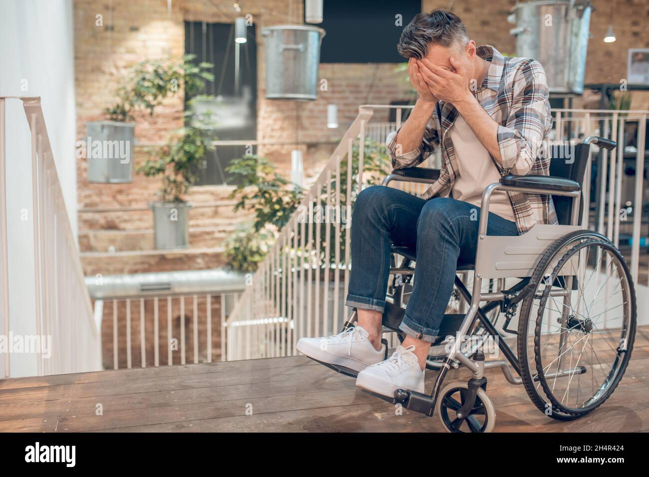 Uomo in sedia a rotelle che ricopre il viso con le mani Foto Stock