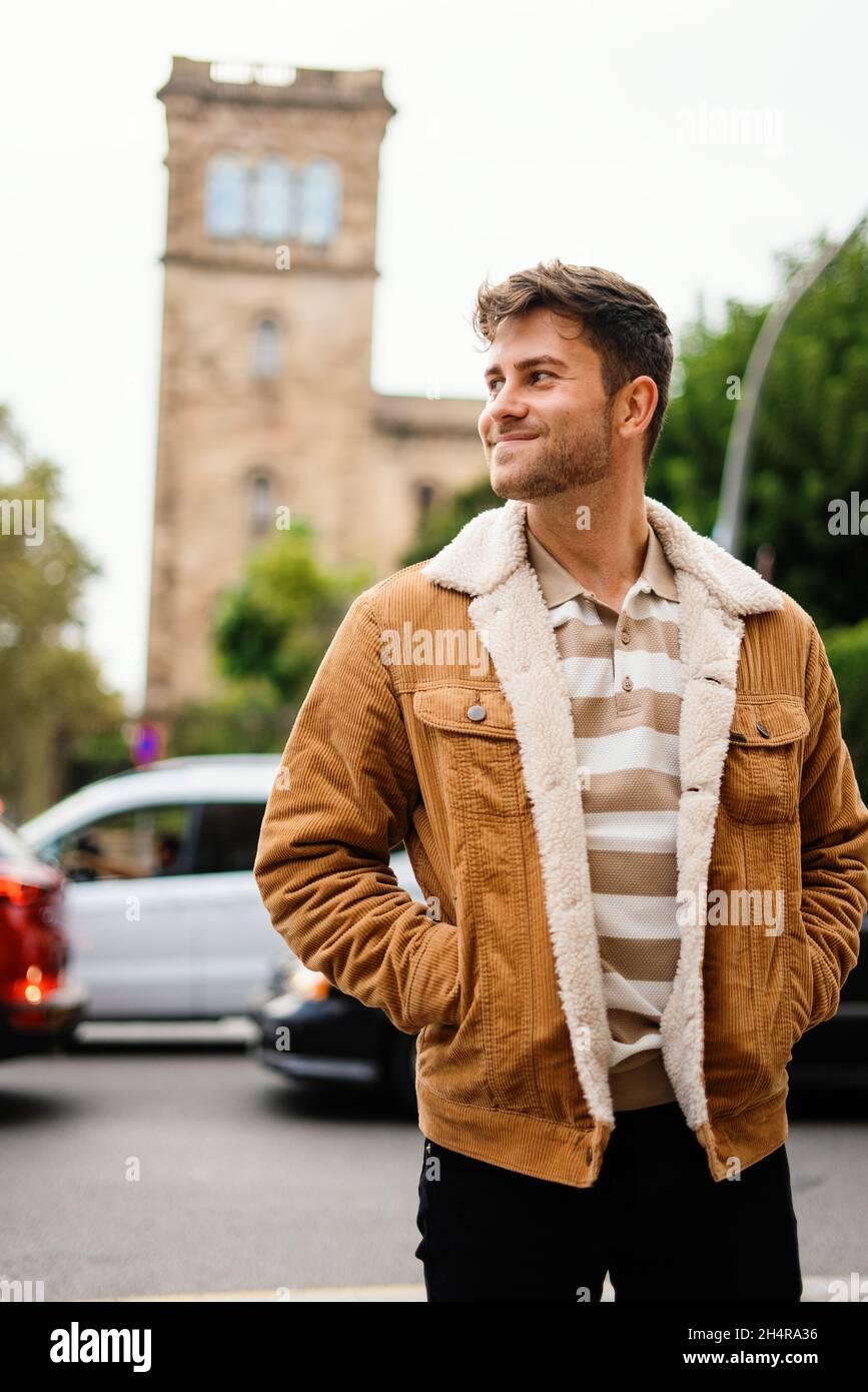 Uomo adulto ottimista che tiene le mani in tasche di giacca calda e  guardare via con sorriso mentre si sta in piedi vicino alla strada con le  automobili sulla strada della città