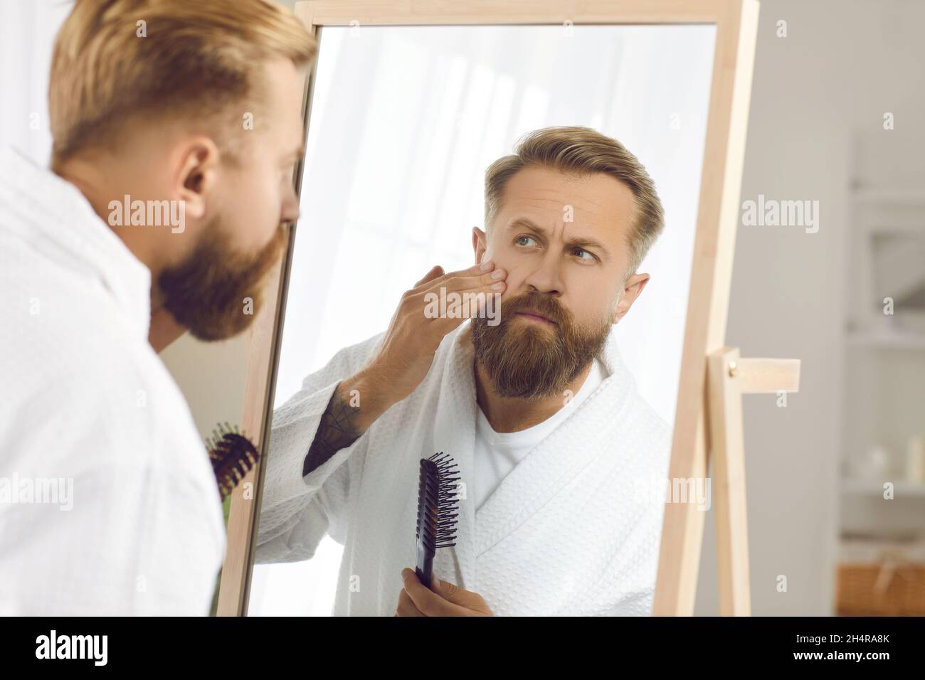 Uomo preoccupato che guarda le rughe sul viso nello specchio durante la routine di cura della pelle Foto Stock