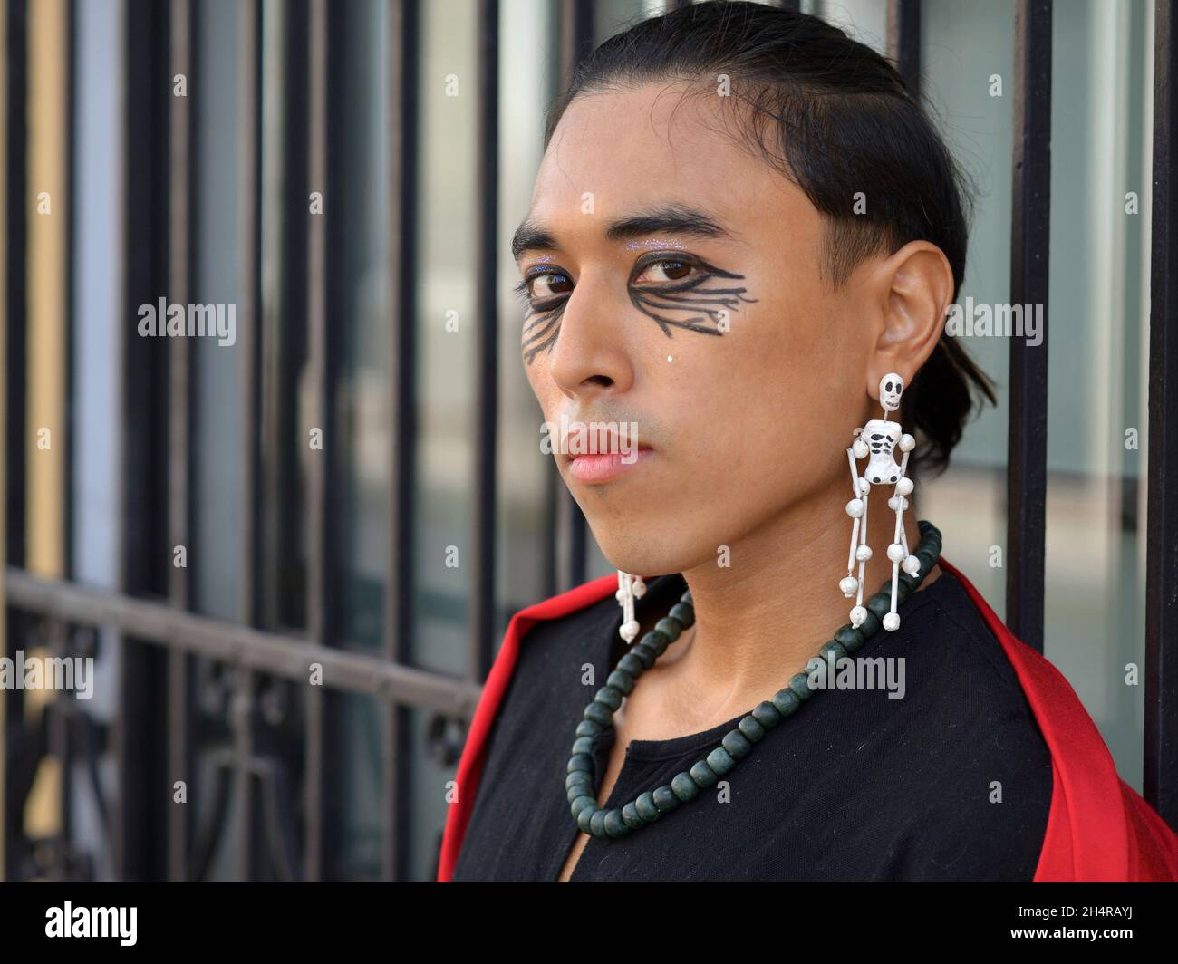 Il bel modello messicano transgender non binario con gli orecchini a scheletro Day of the Dead guarda lo spettatore davanti alle barre nere del burglar. Foto Stock