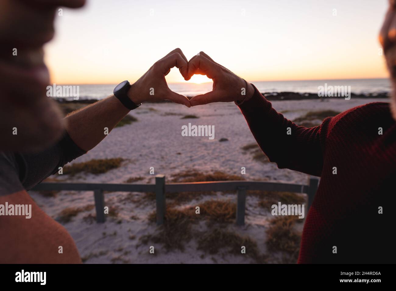 Felice coppia maschio gay caucasico che forma la forma del cuore con le mani sulla spiaggia al tramonto Foto Stock