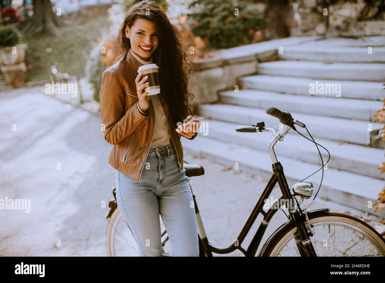 Bella giovane donna con cellulare bere caffè per andare in bicicletta il giorno d'autunno Foto Stock