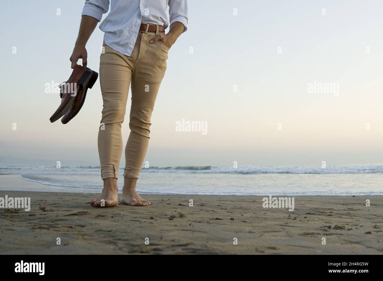 Uomo elegante di classe in camicia bianca e pantaloni beige che cammina  sulla spiaggia con le scarpe in mano Foto stock - Alamy