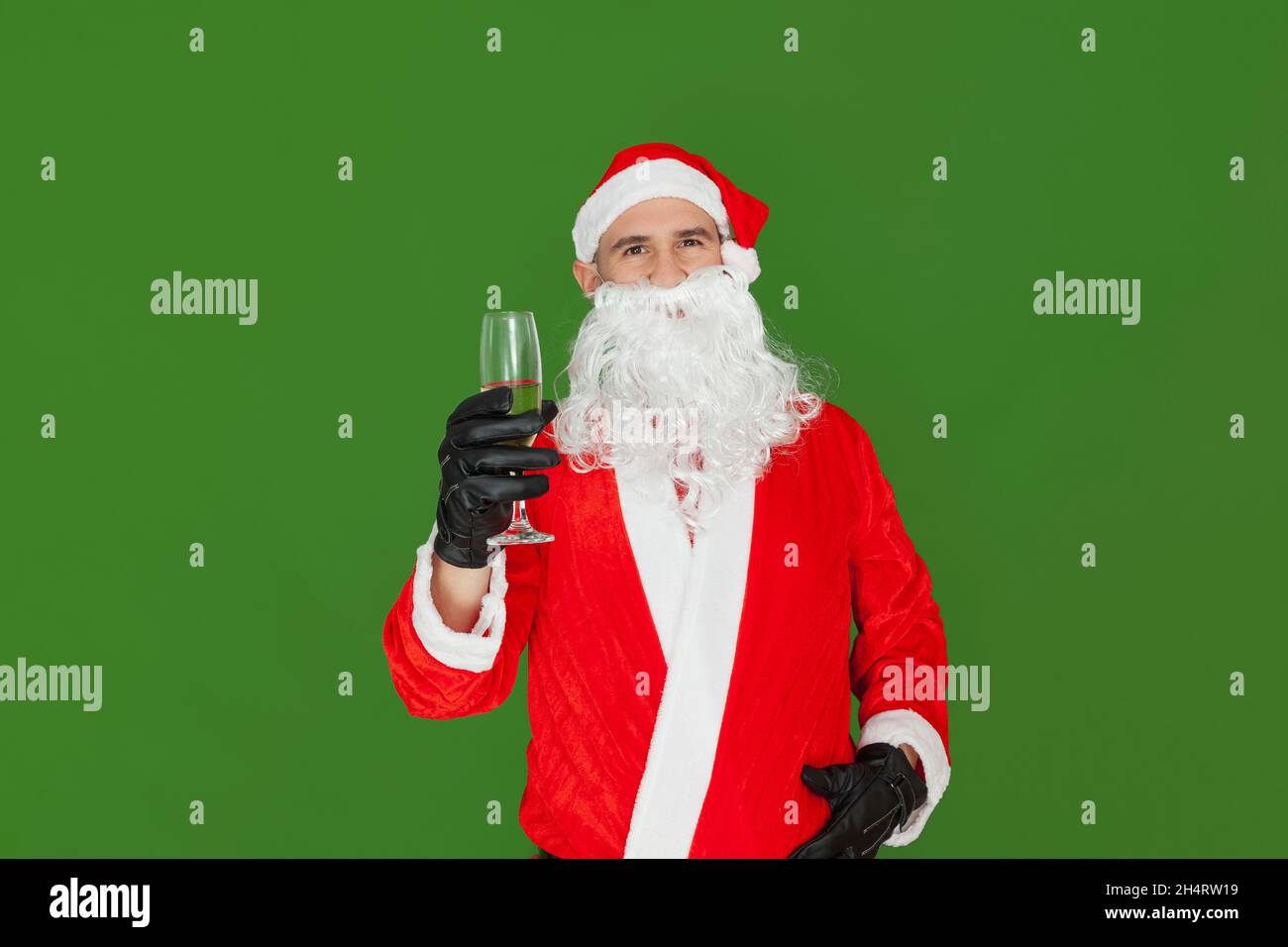 Un uomo caucasico vestito come Babbo Natale sta tenendo una tazza di vetro piena di champagne nella sua mano destra mentre guarda verso la macchina fotografica. Lo sfondo è Foto Stock