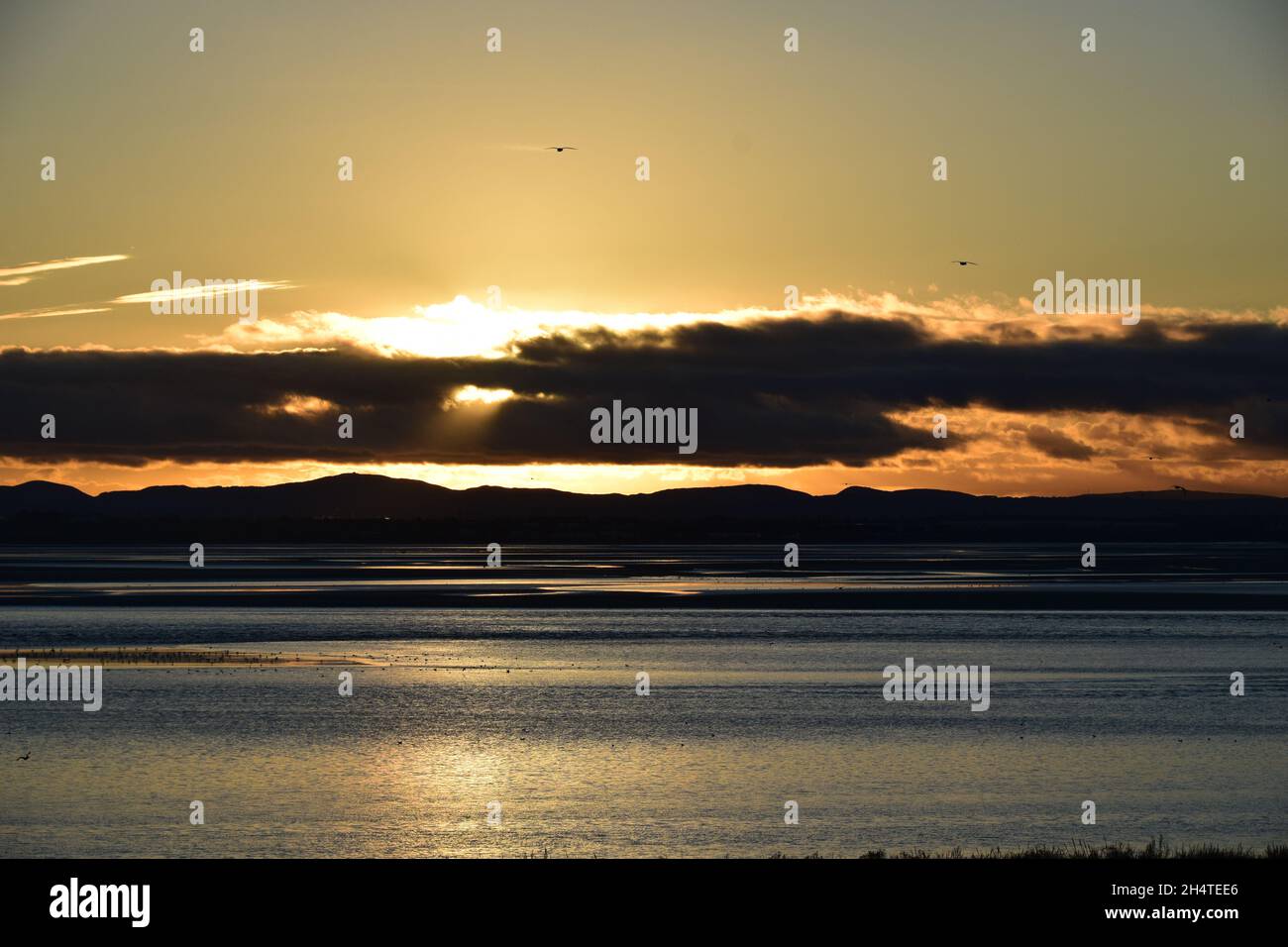Il sole tramonta verso il basso rivelando riflessi d'acqua scintillanti e nuvole scure sopra l'estuario del fiume Mersey. Foto Stock