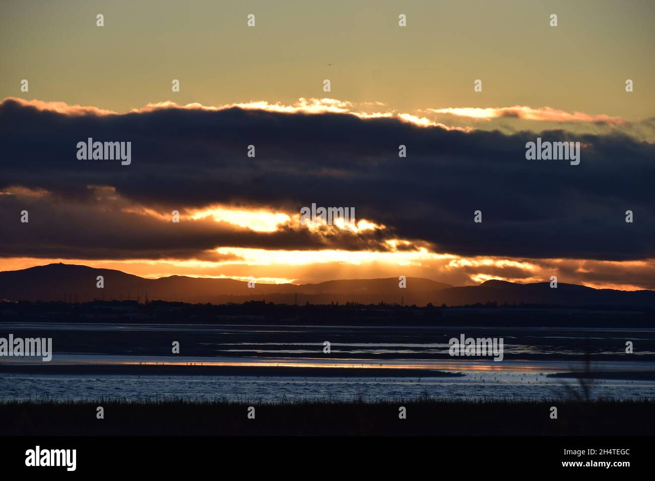 Il sole tramonta verso il basso rivelando riflessi d'acqua scintillanti e nuvole scure sopra l'estuario del fiume Mersey. Foto Stock