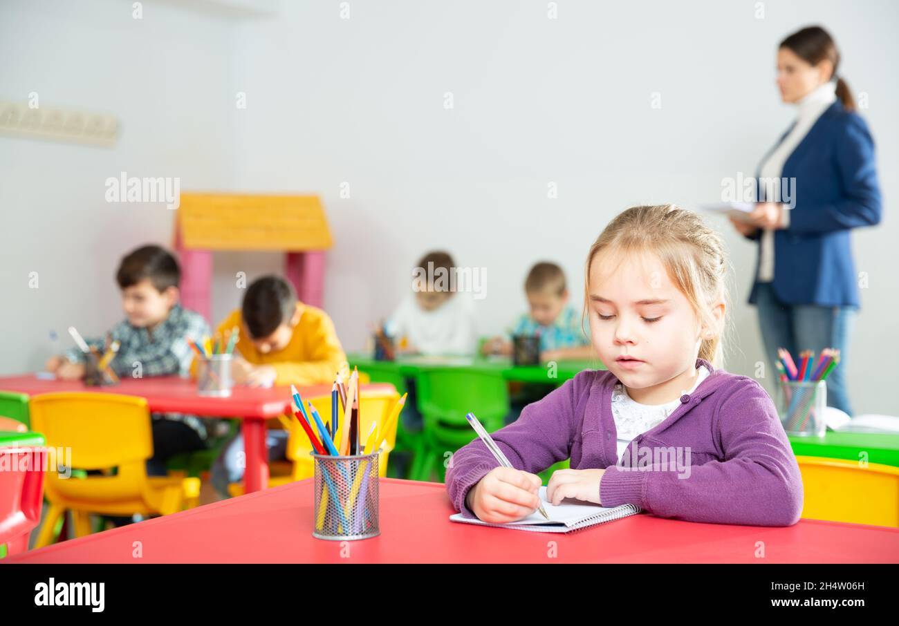 Carino ragazza di età che studia in classe su sfondo con compagni di classe e insegnante Foto Stock