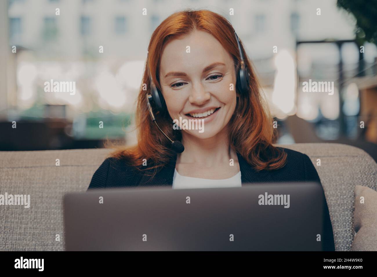 Sorridente felice giovane donna rossa che indossa le cuffie parlando con i colleghi online su un laptop Foto Stock