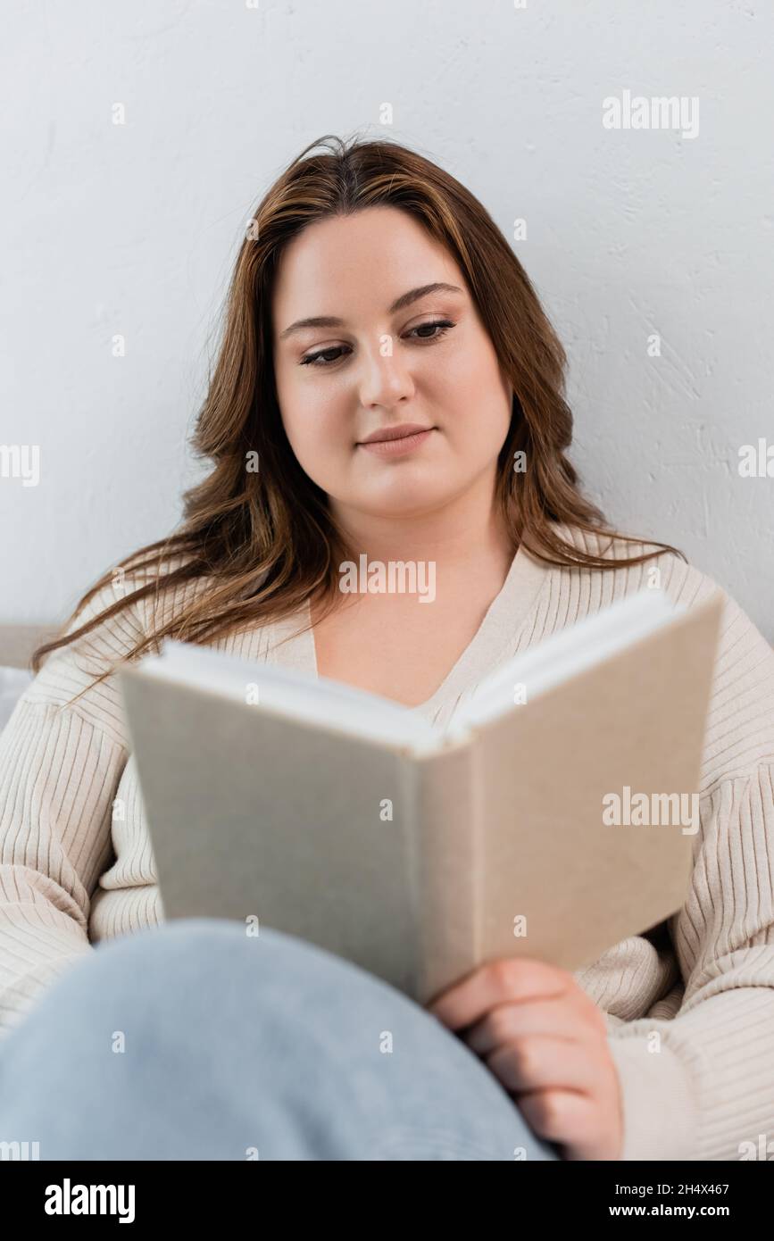 Donna brunetta con lettura in sovrappeso offuscato libro a casa Foto Stock