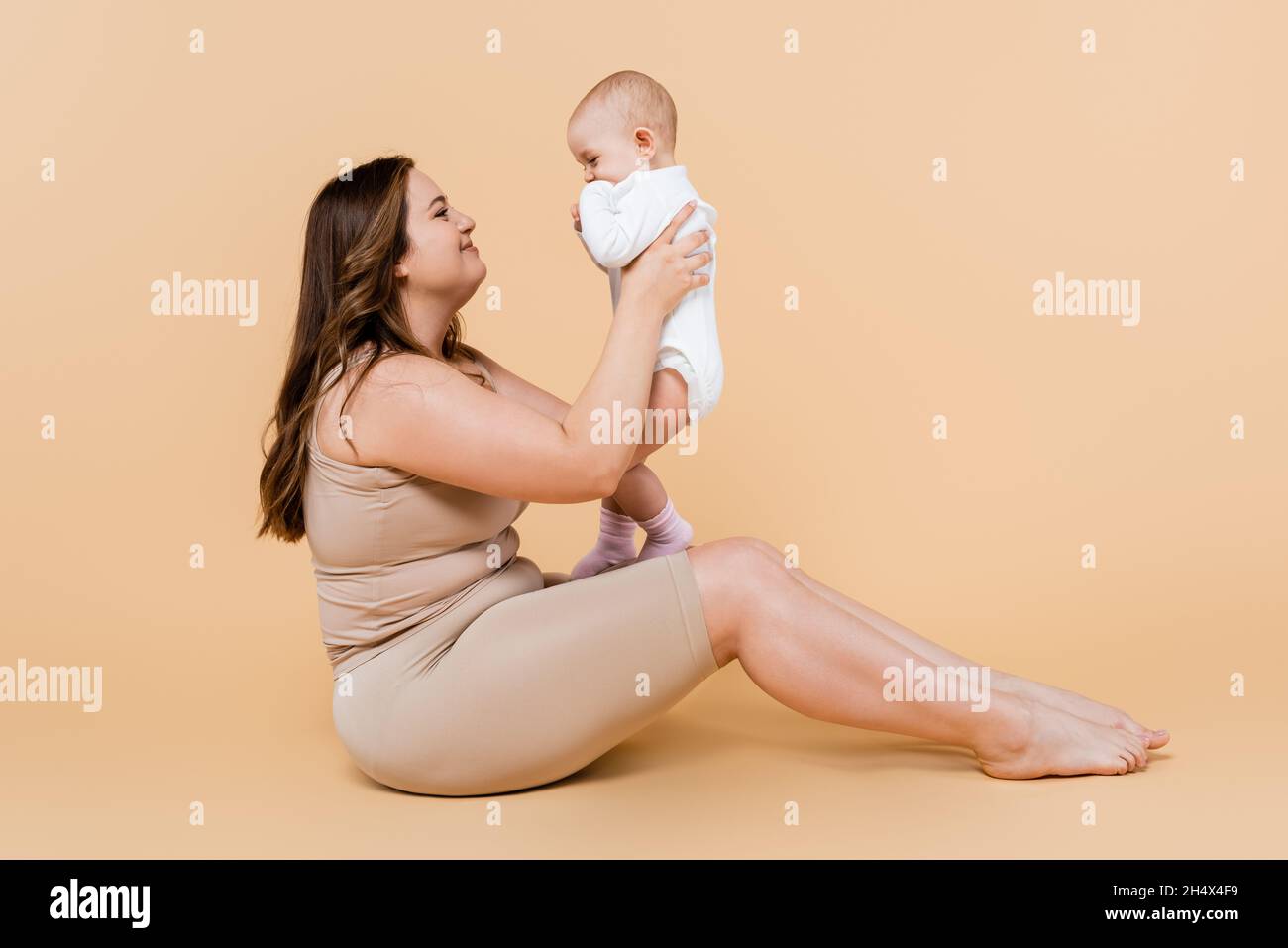 Vista laterale di una madre sorridente più grande che tiene il bambino mentre si siede su sfondo beige Foto Stock