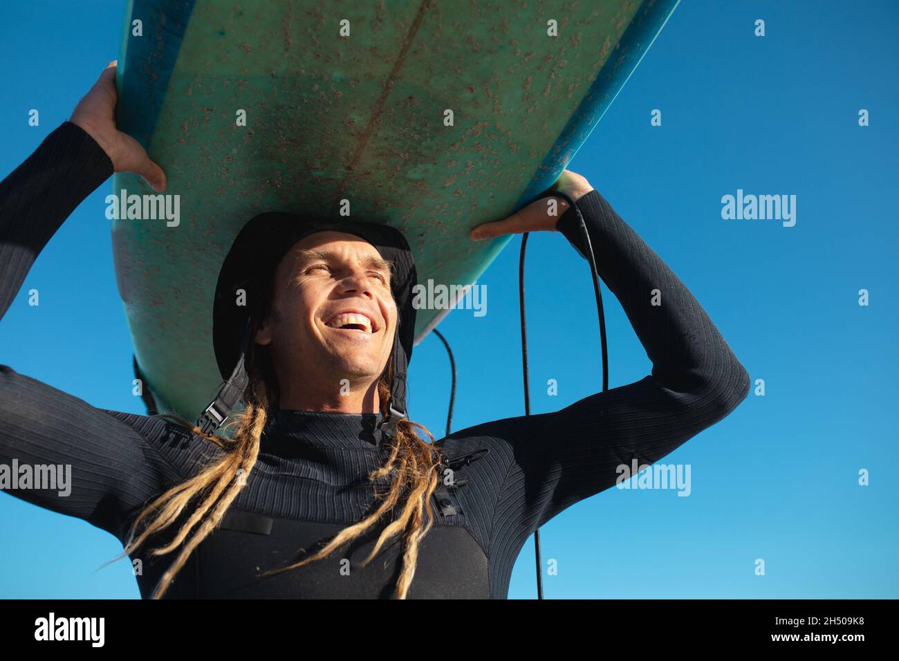 Felice surfista maschile portando surf in testa contro cielo blu chiaro in giorno di sole Foto Stock