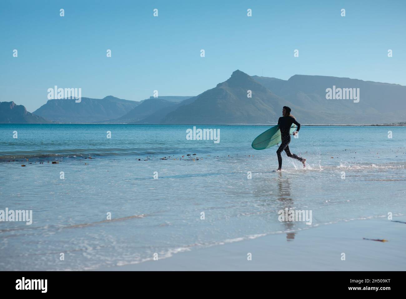 Surfer maschio che trasporta la tavola da surf mentre corre su terra spruzzi d'acqua contro lo spazio di copia Foto Stock