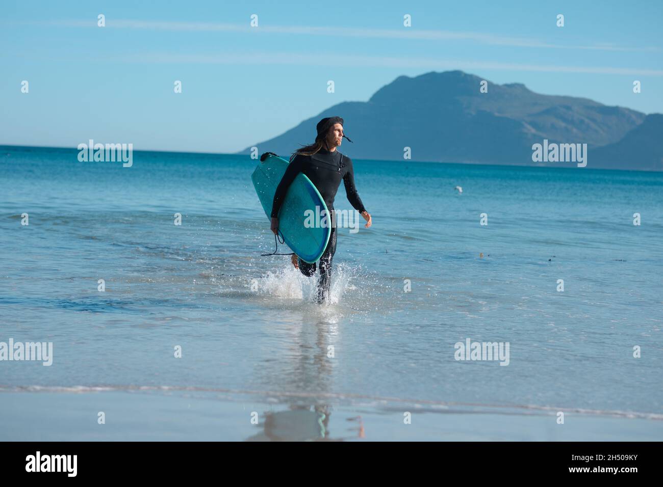 Uomo con tavola da surf che corre mentre spruzzi d'acqua a riva in spiaggia durante la giornata di sole Foto Stock