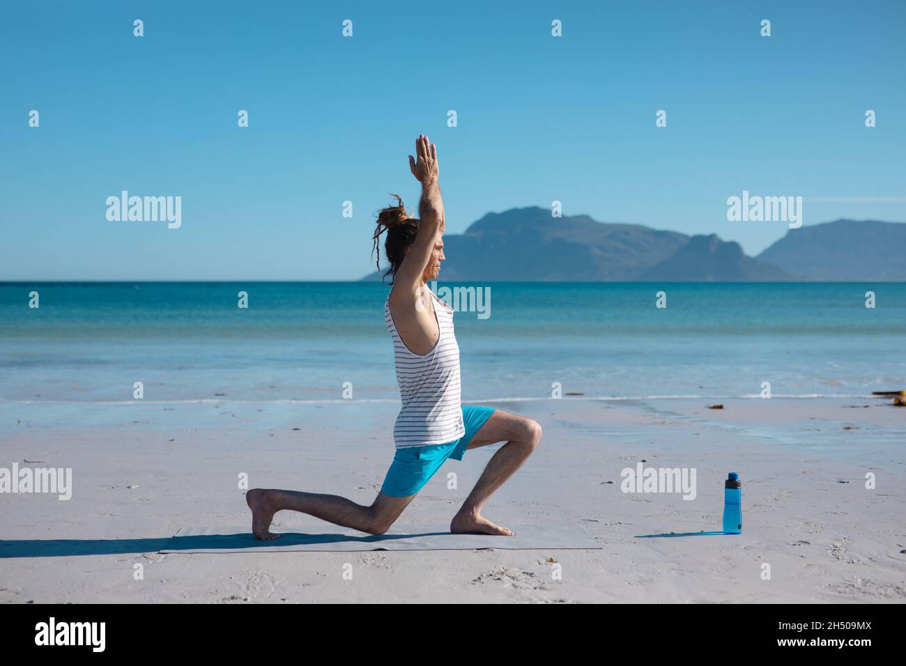 Vista laterale dell'uomo che pratica il guerriero 1 posa yoga con le mani aggrappate sulla testa alla spiaggia, spazio copia Foto Stock