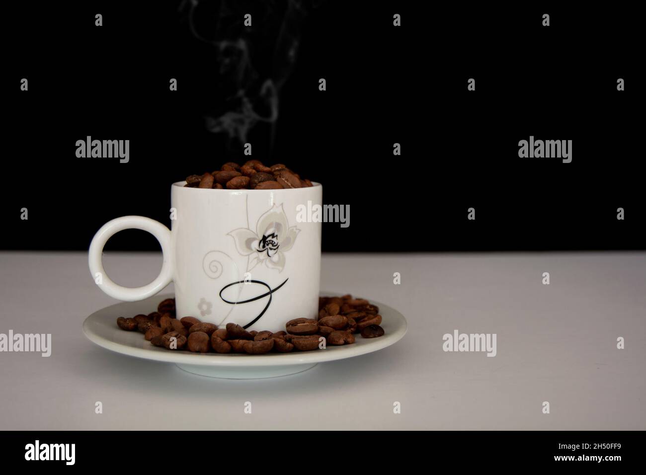 Caffè caldo in una tazza di caffè bianca e molti chicchi di caffè posti su un tavolo bianco in un'atmosfera calda e chiara, su sfondo scuro, con spazio per la copia. Foto Stock