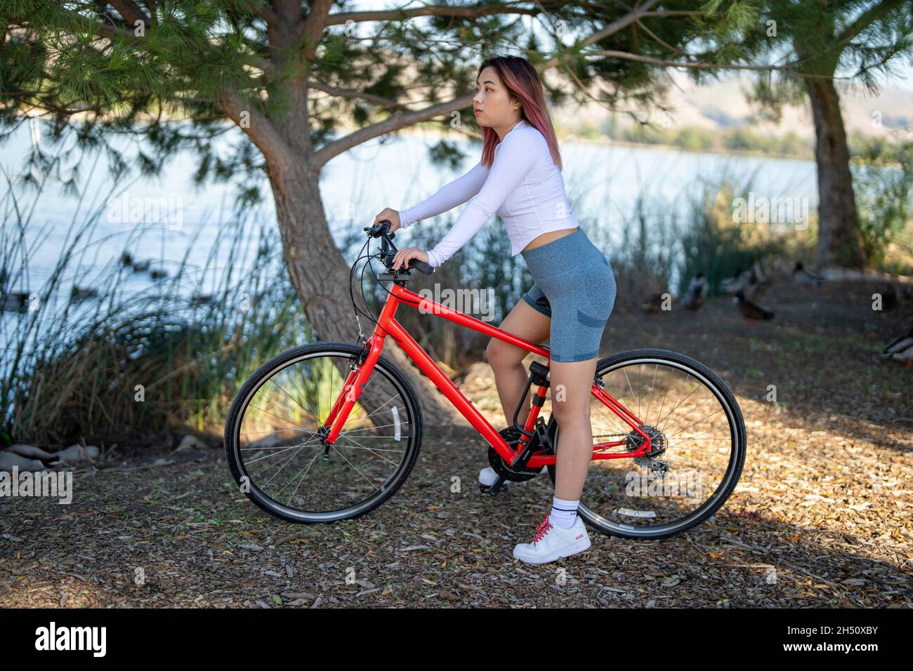 Giovane donna asiatica seduta su una bicicletta ibrida arancione di fronte ad un lago Foto Stock