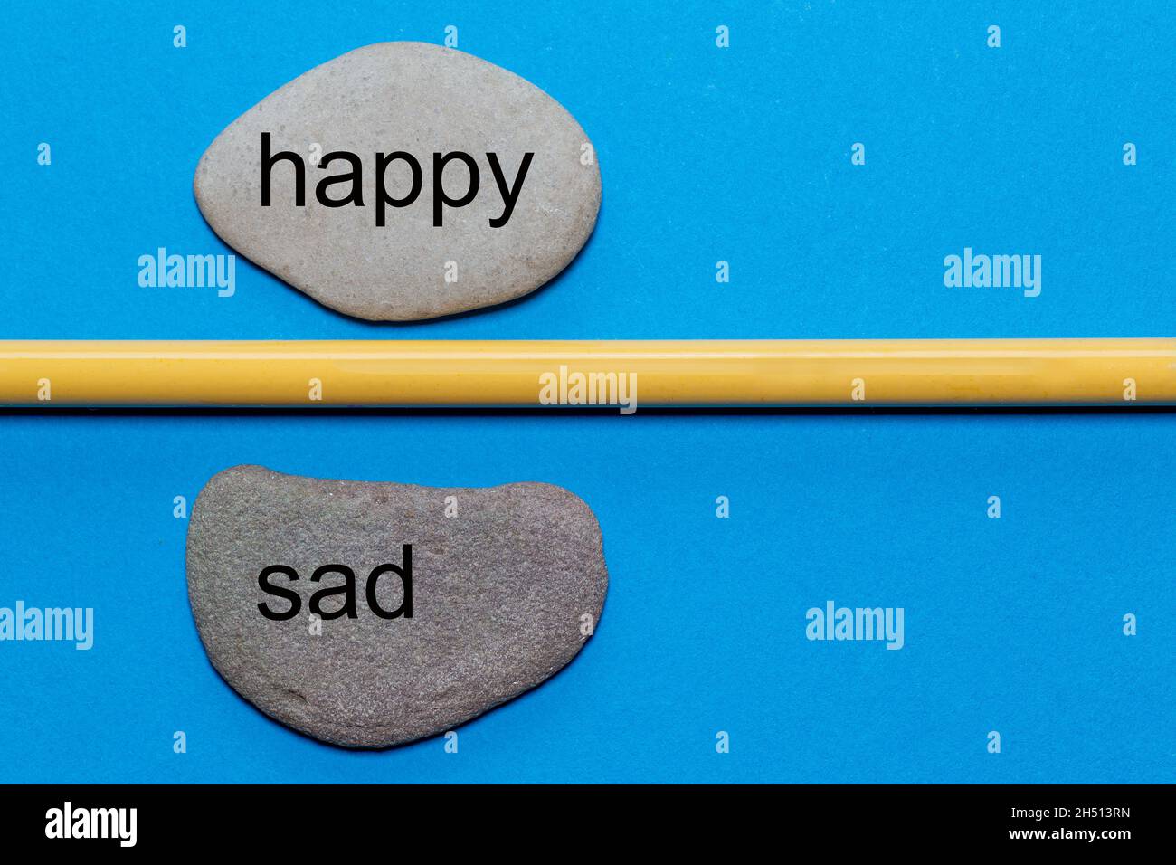 Le parole felice e triste sono scritte su pietre naturali lisce separate da una matita gialla. Lo sfondo è isolato in blu e ha molto spazio Foto Stock