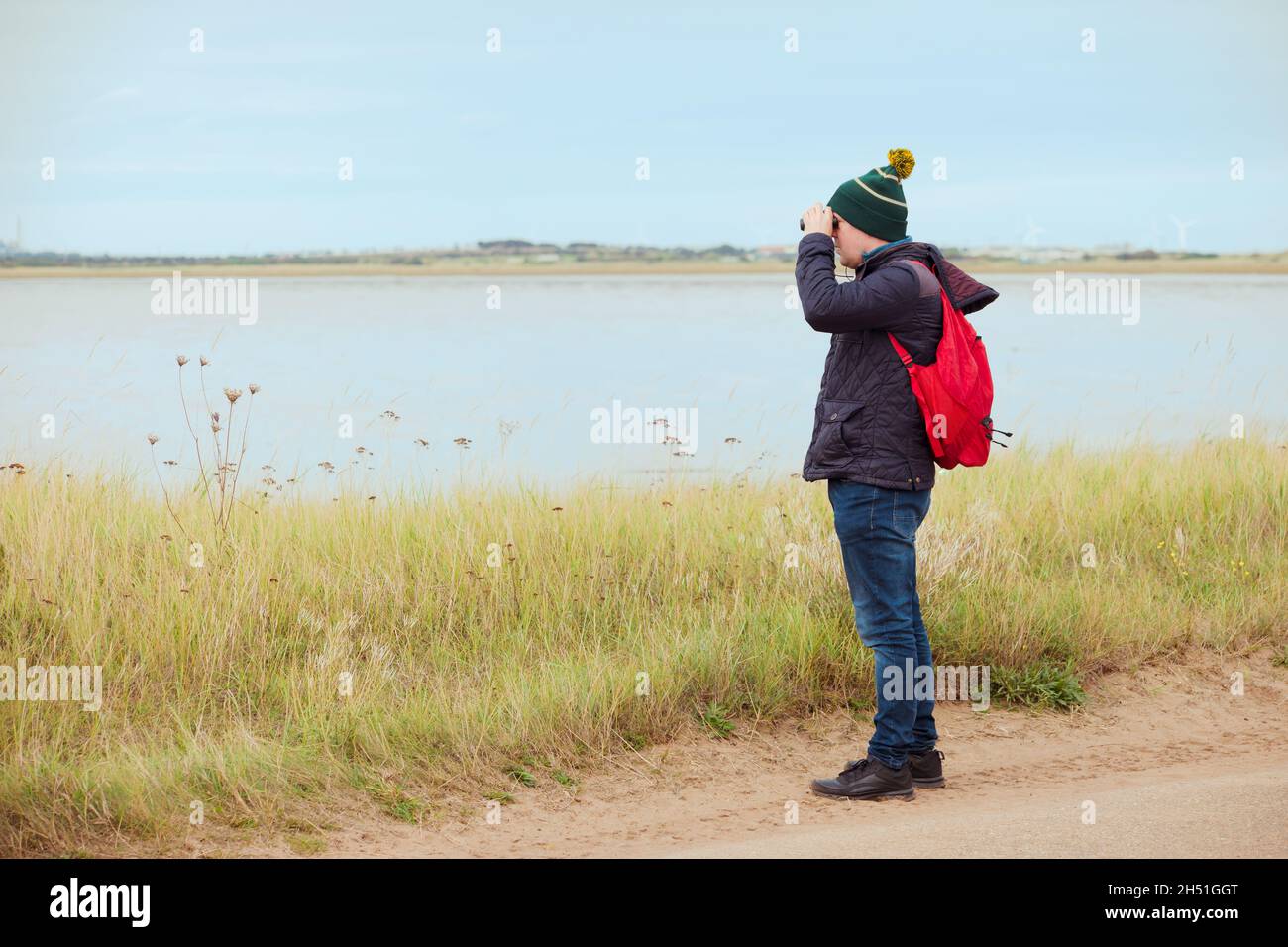 Un birdwatcher maschio adulto in cappotto caldo e cappello con binocolo e zaino in mare o lago Foto Stock