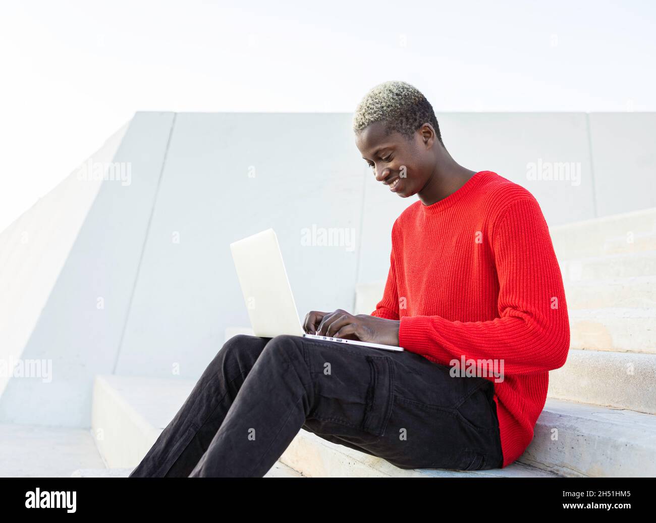 Ragazzo africano nero con computer portatile. Felice e sorridente. Seduto su scale su sfondo bianco. Foto Stock