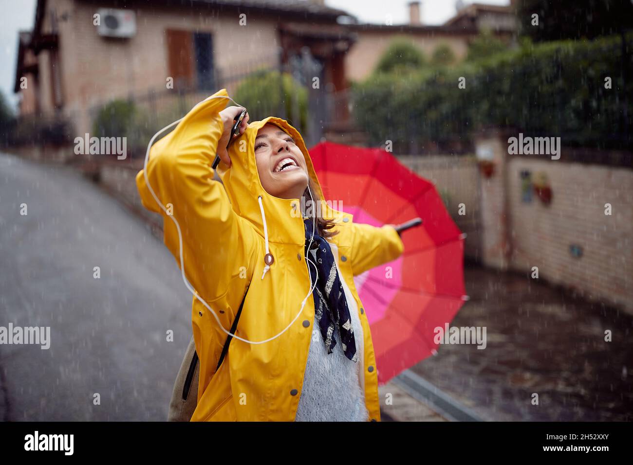 Una ragazza giovane in un impermeabile giallo e con ombrello rosa che cammina per la strada in una giornata nuvolosa è di buon umore mentre si gode la musica e la pioggia dentro Foto Stock