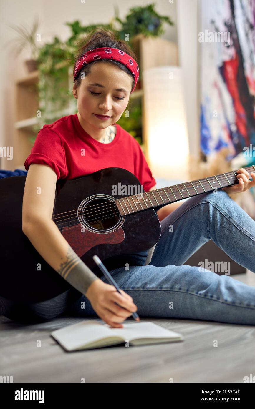 Giovane donna creativa in abbigliamento casual che fa appunti nel suo notebook mentre suona la chitarra acustica, studiando musica a casa Foto Stock