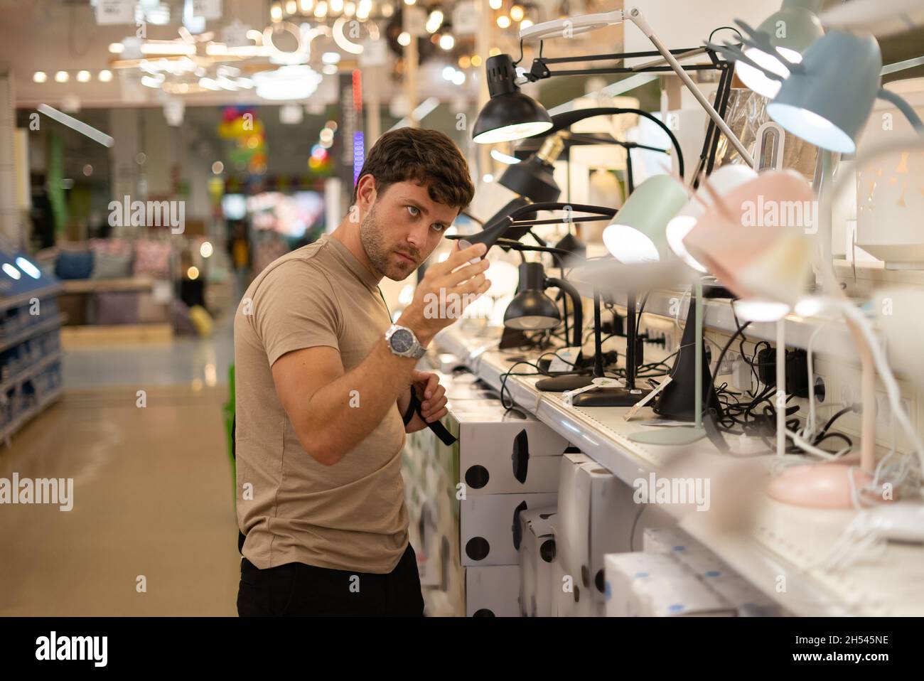Il tipo pensivo controlla la lampada da tavolo mentre acquista nel negozio moderno con arredamento domestico Foto Stock