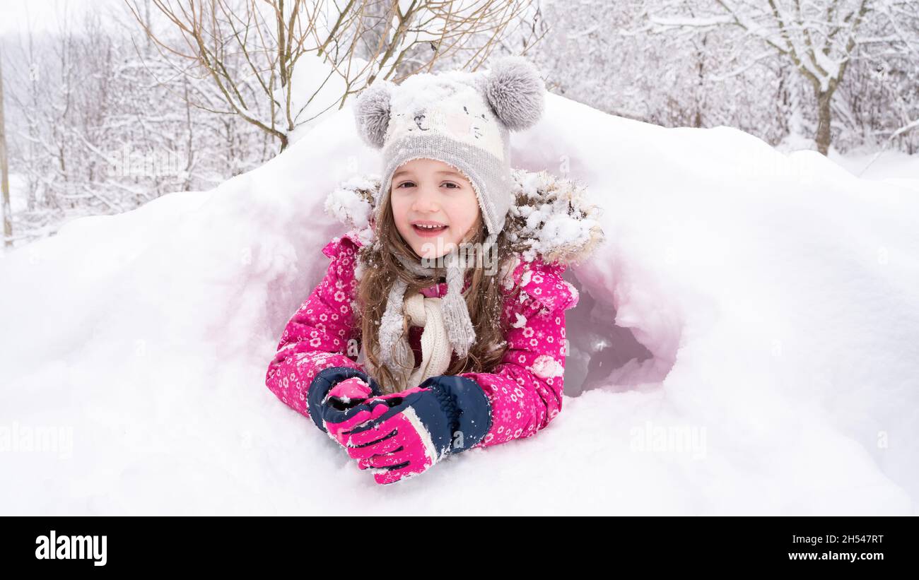La ragazza si trova in una nevicata e sorride. Il bambino in un cappello divertente ha costruito un igloo o una grotta di neve. Idee per l'attività invernale per i bambini. Cosa fare per Foto Stock