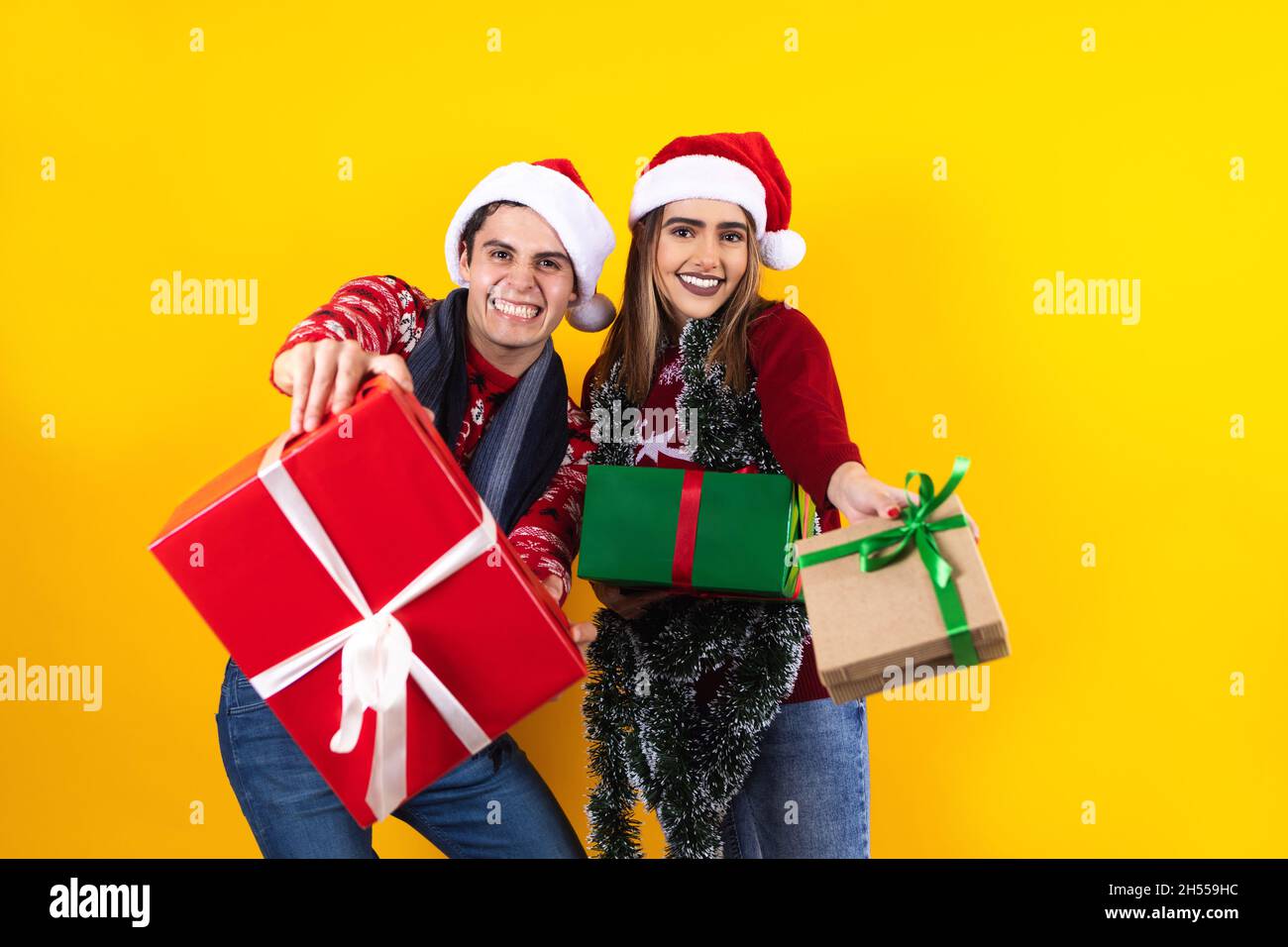Giovane Santa latin coppia amici uomo e donna in rosso maglione cappello di Natale tenere presente scatola con regalo fiocco nastro su sfondo giallo in Messico latino Foto Stock