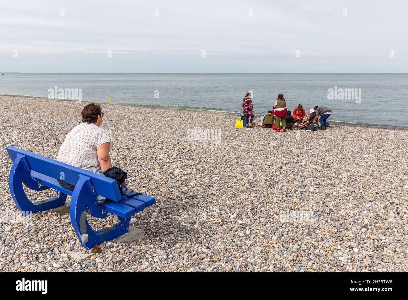 Signora seduto su una panca pubblica blu sulla spiaggia di ciottoli di le Tréport, osservando un gruppo di persone al bordo dell'acqua. Le Tréport, Francia Foto Stock
