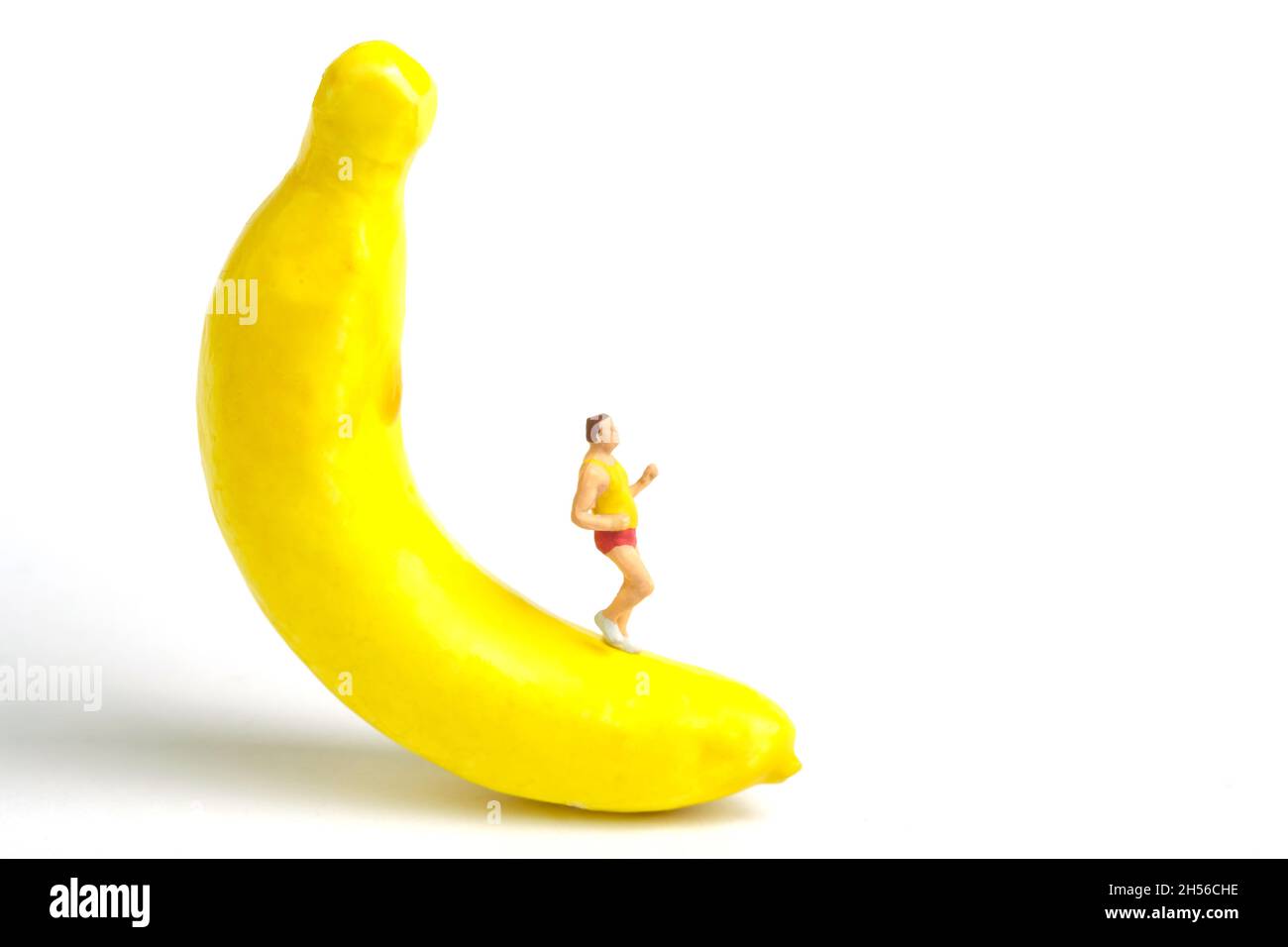 Fotografia di personaggi giocattolo in miniatura. Concetto di combustione dei grassi. Un uomo grasso che corre al di sopra della banana frutta, isolato su sfondo bianco. Foto immagine Foto Stock