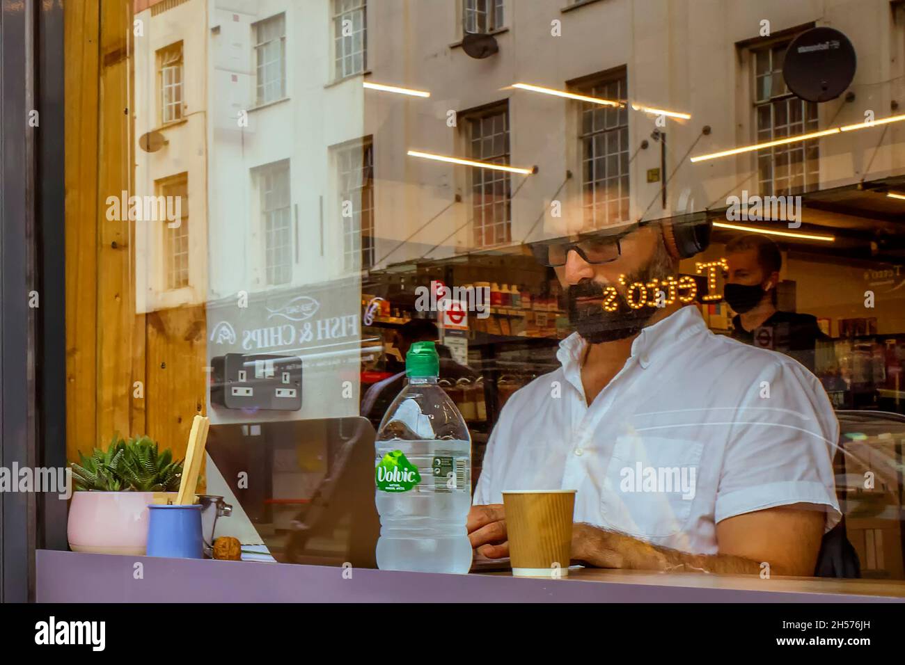 Attraverso la finestra di una caffetteria si può vedere un uomo che si concentra e indossa le cuffie mentre si lavora su un computer portatile. Foto Stock
