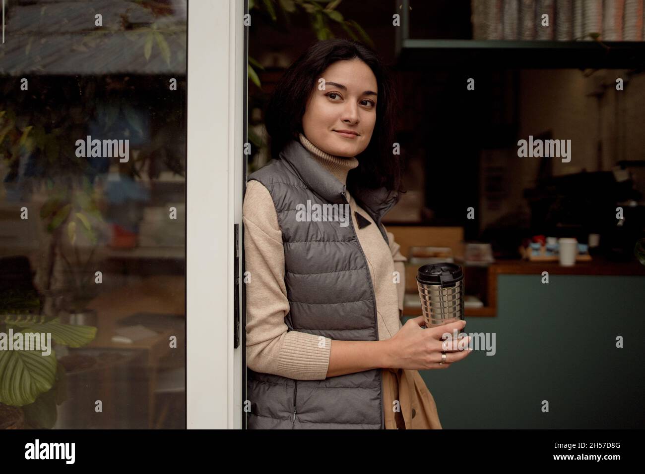 Giovane donna in outerwear gustando una bevanda calda e guardando via mentre si appoggia alla porta del caffè al mattino Foto Stock