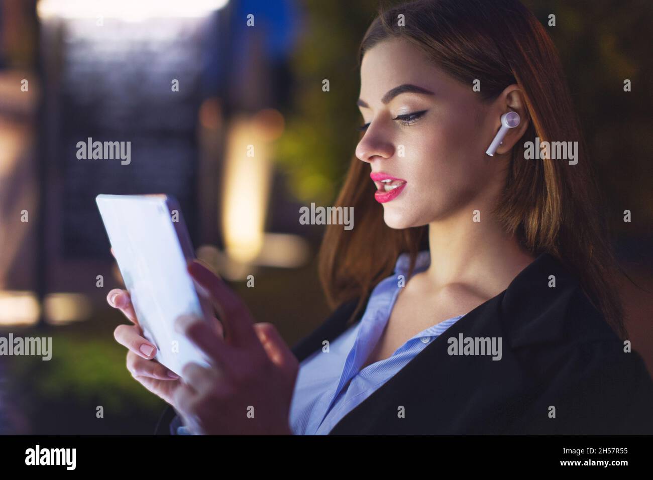 Giovane donna d'affari con dispositivo vivavoce che utilizza il tablet di notte in città, chiamata vocale Foto Stock