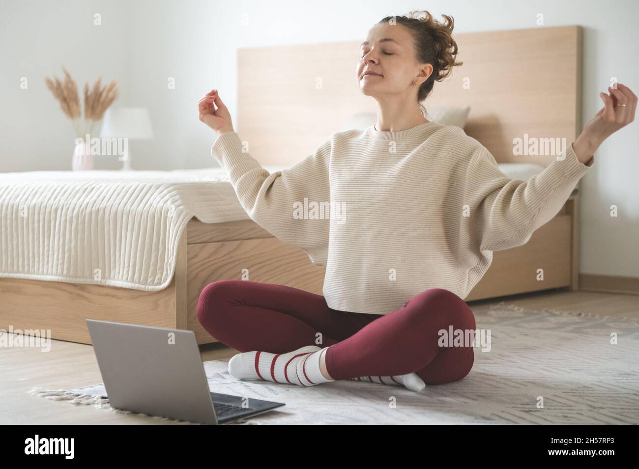 Donna piuttosto sportiva che fa yoga o pilates in camera da letto accogliente a casa utilizzando lezione virtuale con il computer portatile Foto Stock