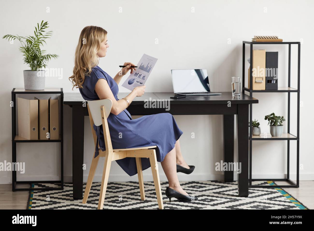 Ritratto a tutta lunghezza di elegante donna d'affari che legge i documenti dalla scrivania in minimo ufficio interno, spazio di copia Foto Stock