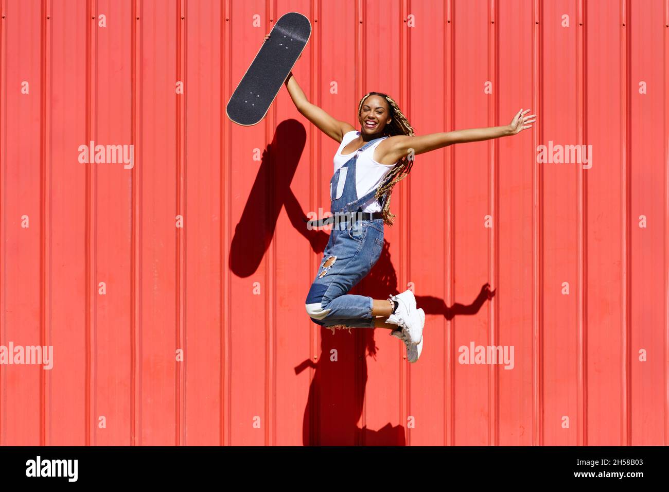 Donna nera vestita casual, wtih uno skateboard saltando con felicità su sfondo rosso muro urbano. Foto Stock
