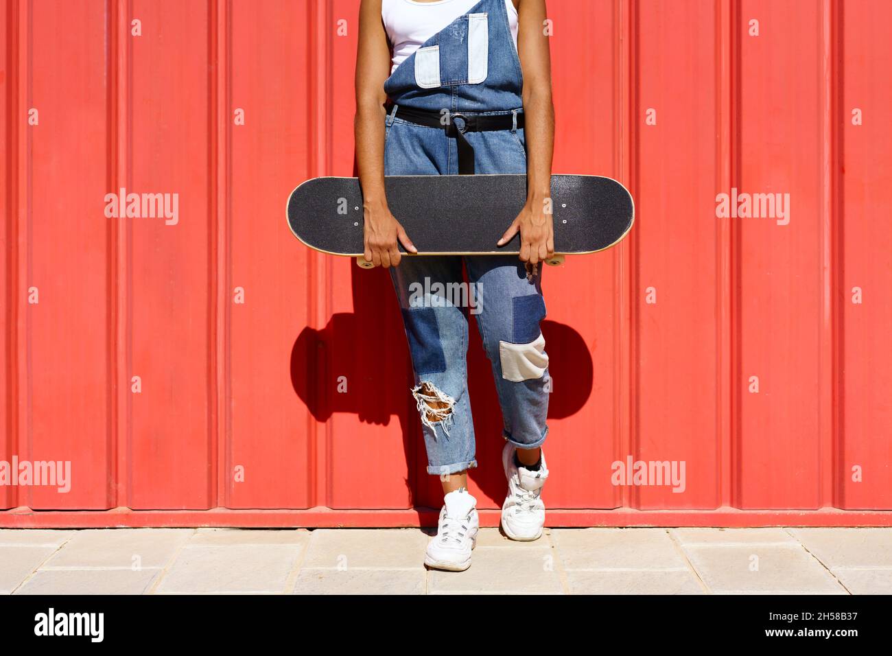 Donna irriconoscibile che tiene uno skateboard con le mani su un muro urbano rosso. Foto Stock