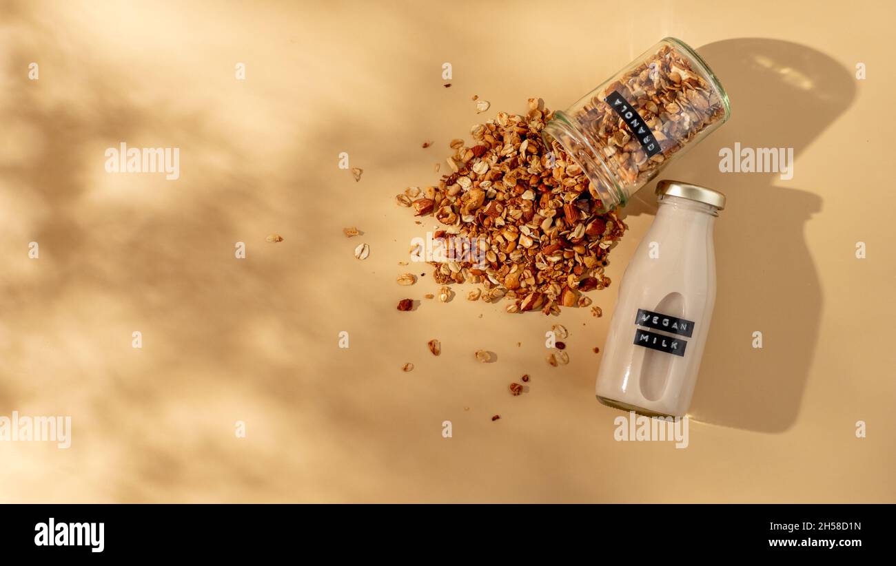 Granola e latte vegano. Colazione salutare concetto estetico. Granola fatta in casa cosparso di vasetto di vetro e latte vegano in bottiglia di vetro su sfondo giallo champagne o beige pastello. Vista dall'alto, disposizione piatta Foto Stock