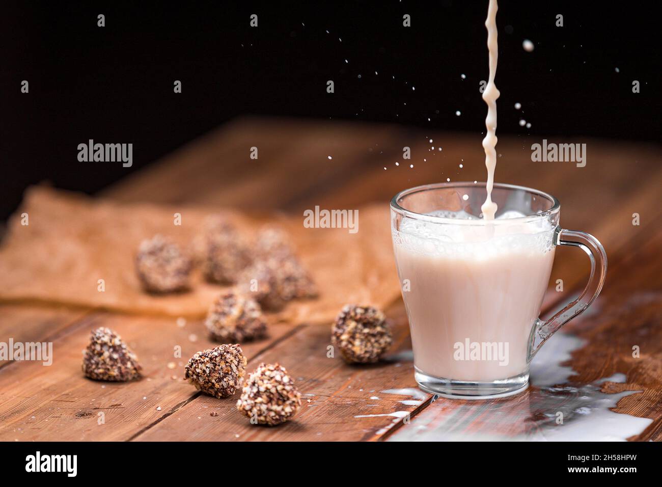 Un getto di latte si versa in una tazza di vetro di tè, formando uno spruzzo. Il latte è versato su un tavolo di legno. Dolci sul tavolo. Foto Stock