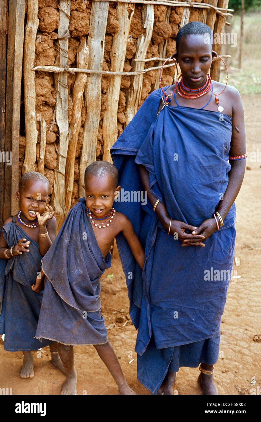 Africa, Tanzania, 1976. Ujamaa villaggio Maasai persone ancora vivono nelle loro case tradizionali. Maasai donna e due dei suoi figli in piedi di fronte alla loro capanna. Foto Stock