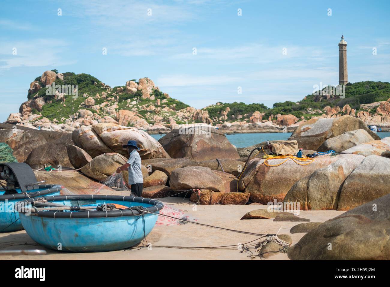 Un pescatore locale svella la rete di pesca sulla riva del mare. Spiaggia pittoresca con grandi rocce. Vecchio faro si trova in lontananza. Alta qualità Foto Stock