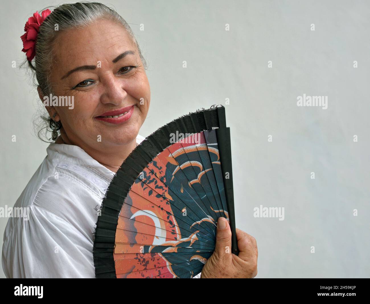 Sorridente elegante donna messicana matura latina con fiore rosso in capelli grigi tiene il ventilatore asiatico portatile e guarda il visore di fronte al muro grigio. Foto Stock