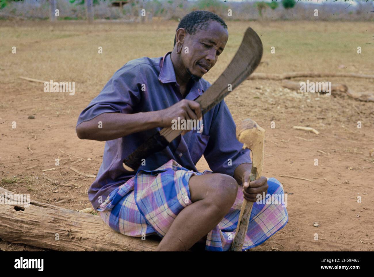 Africa, Tanzania, 1976. Ujamaa villaggio Maasai persone ancora vivono nelle loro case tradizionali. Un uomo carves un bastone con un machete. Foto Stock