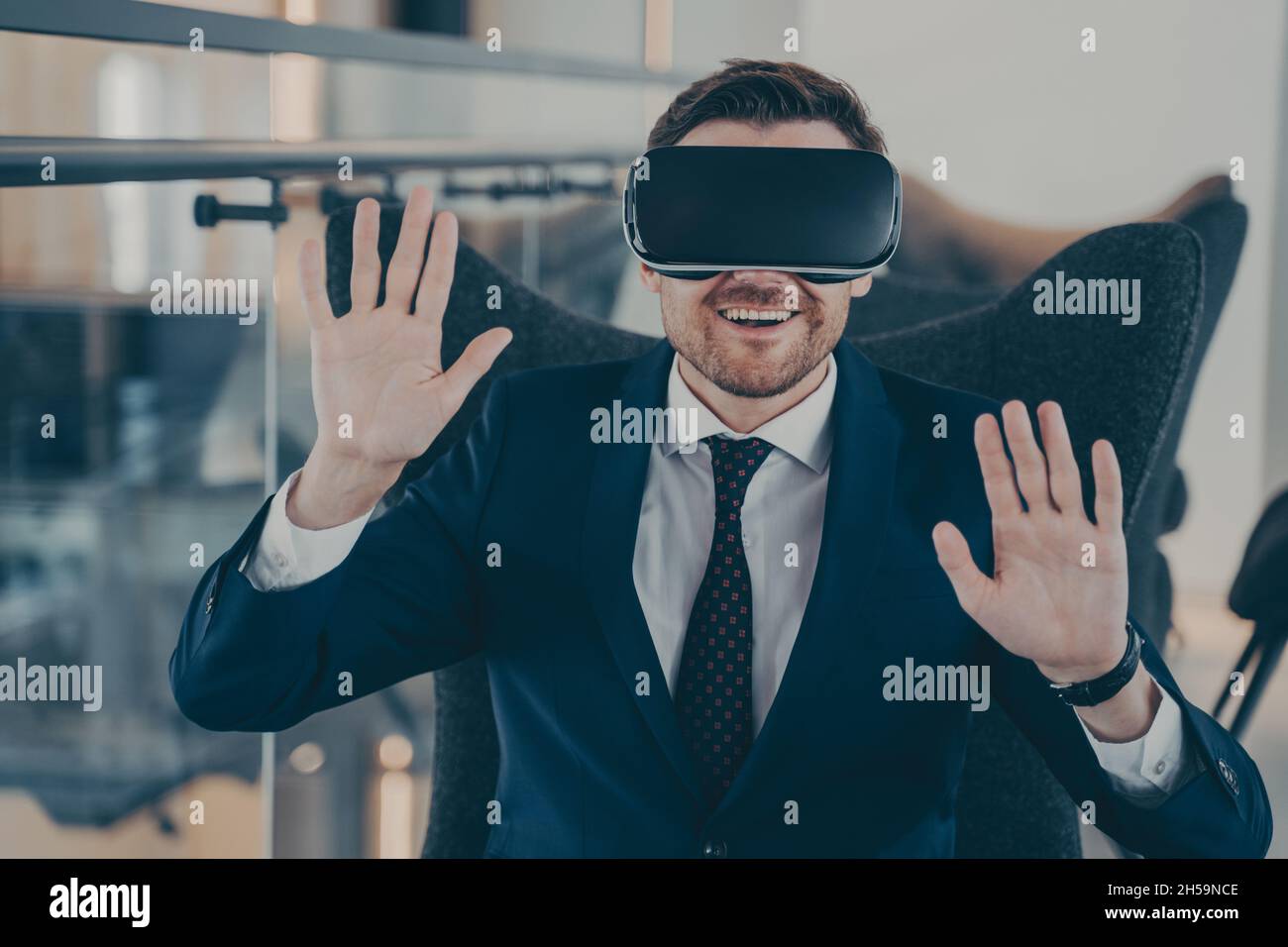 Giovane uomo d'affari sorridente in formale abito nero seduto in ufficio con visore VR Foto Stock