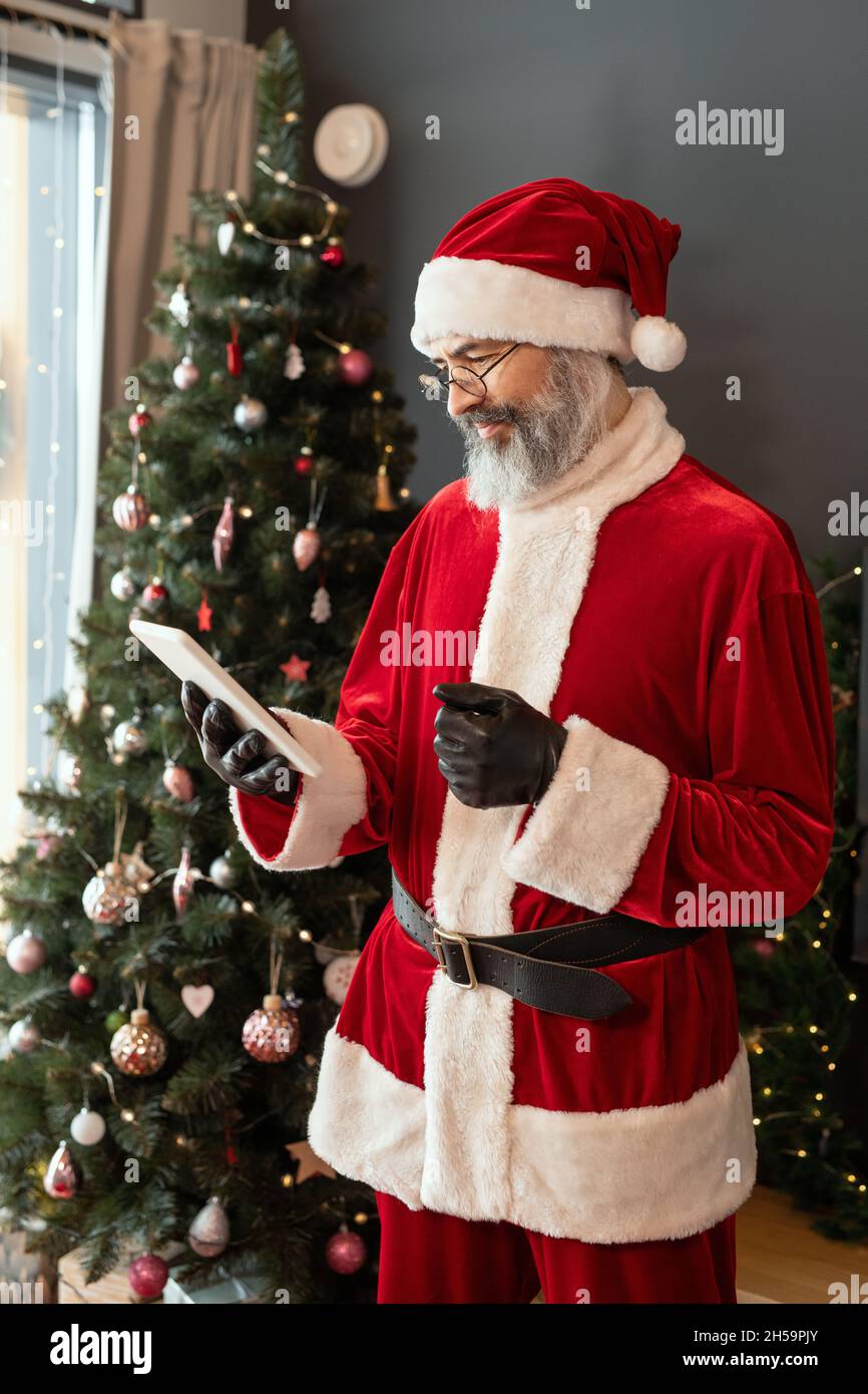 Grave Babbo Natale in costume rosso e occhiali in piedi contro l'albero di Natale e controllare l'e-mail sul tablet Foto Stock