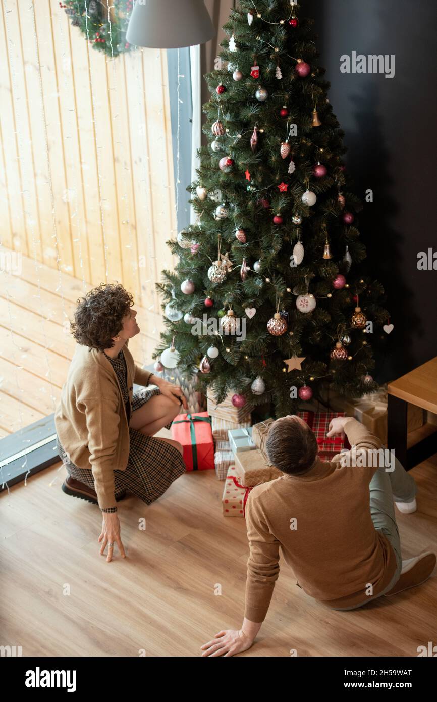 Coppia felice in abiti beige seduta sul pavimento a casa e guardando il bellissimo albero di Natale con giocattoli Foto Stock