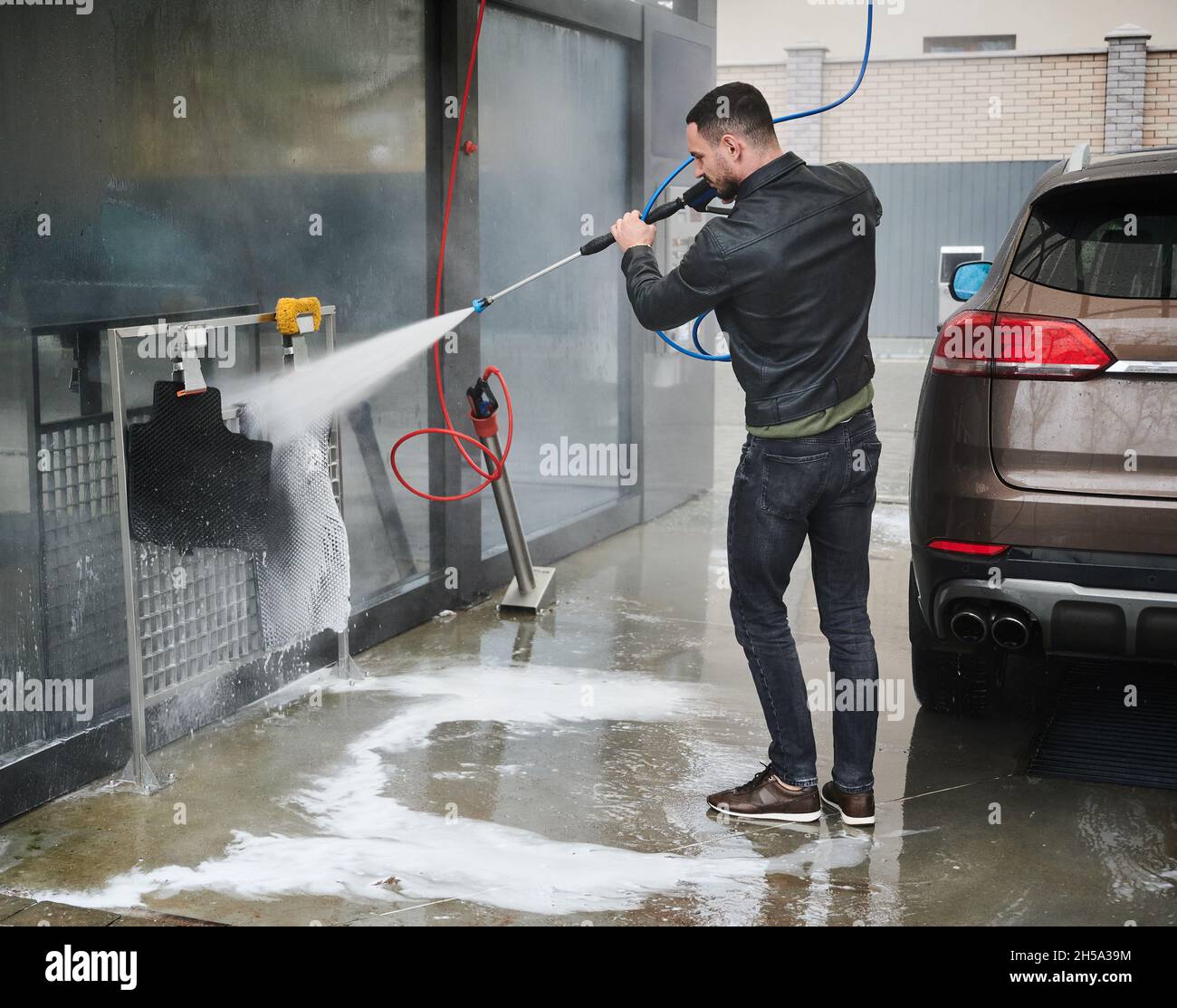 Stazione di lavaggio auto self-service. Giovane conducente maschio in piedi  dietro la sua auto, tenendo in mano l'attrezzatura speciale per il lavaggio  del veicolo. Uomo che utilizza un forte getto di acqua