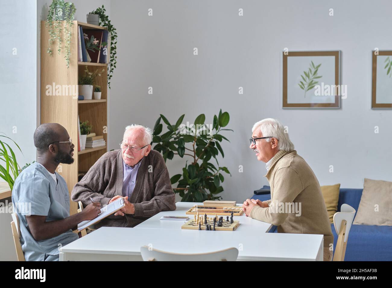Infermiera maschile che dà raccomandazioni agli uomini anziani mentre si siedono al tavolo durante la partita di scacchi in casa di cura Foto Stock
