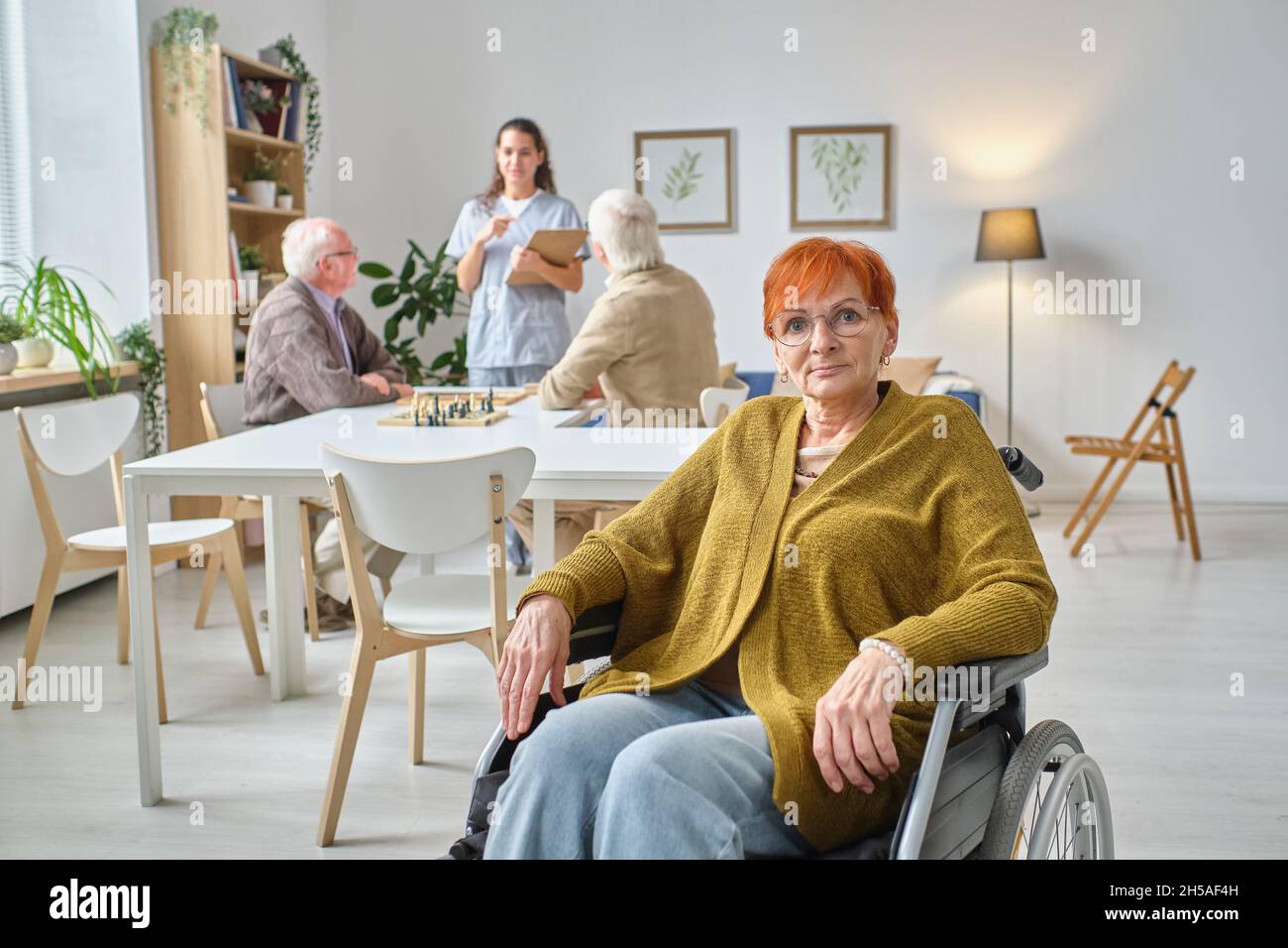 Ritratto di donna matura in occhiali con sedia a rotelle guardando la macchina fotografica seduta nella stanza con altre persone anziane in background Foto Stock